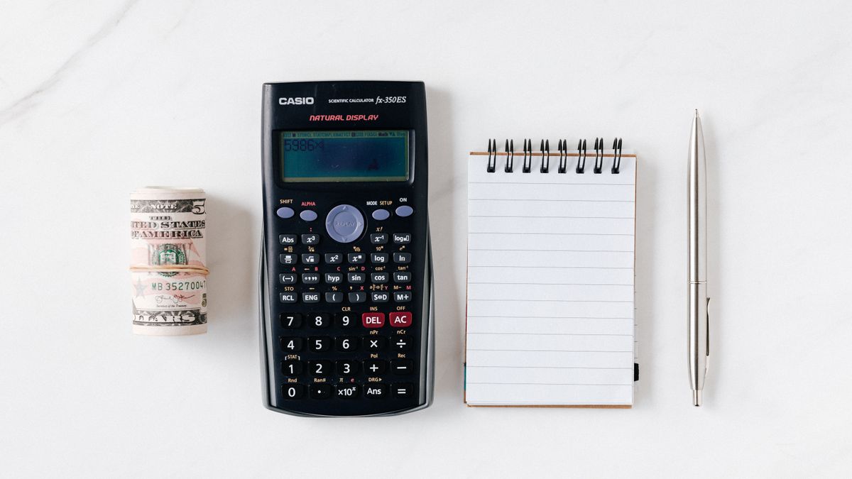 A calculator, a roll of banknotes, a notepad and a pen lined up on a marble surface