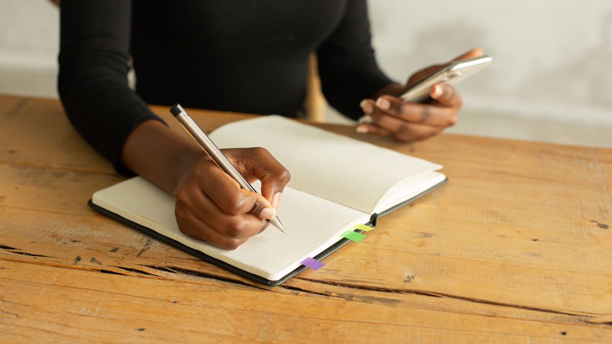 Una donna scrive su un quaderno con una penna mentre tiene il telefono nell'altra mano, seduta a un tavolo di legno