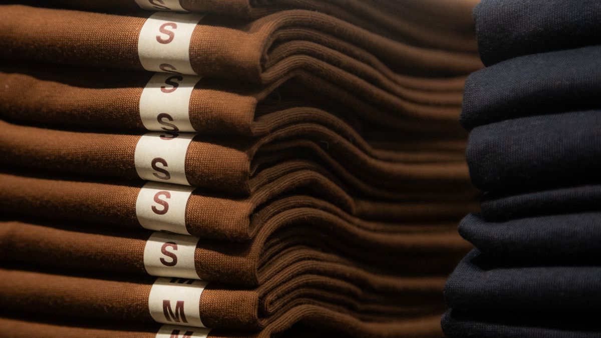 Una pila de camisetas marrones dobladas con las etiquetas de talla a la vista, junto a una pila de camisetas azul marino
